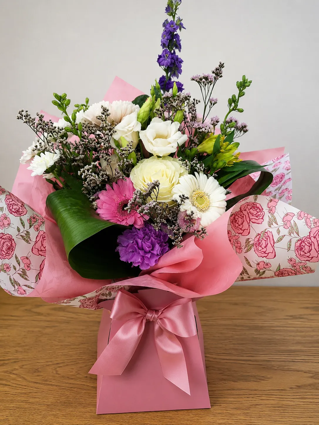 White and purple box arrangement with lisianthus, gerbera and delphinium wrapped in pink rose-print paper