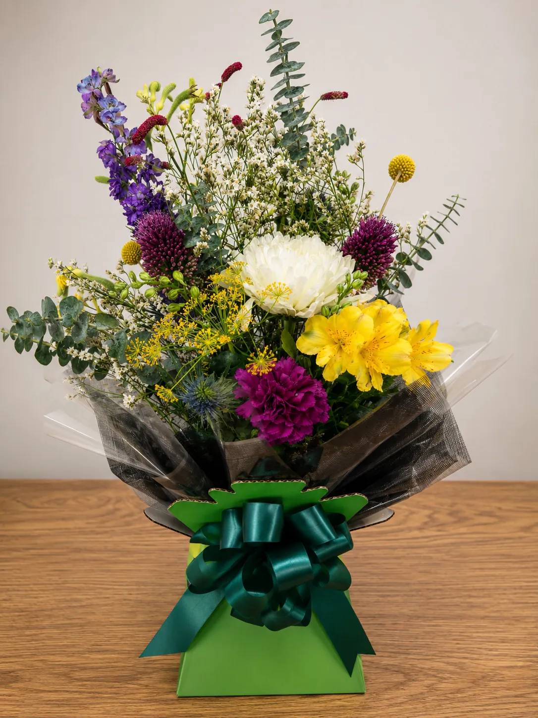 Wildflower-style box arrangement with delphinium, craspedia and blue thistle in a green gift box with a teal bow